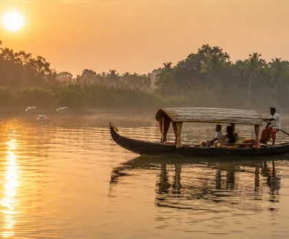 Traditional wooden boat gliding across the calm backwaters during Pathiramanal Island boating in Kerala at sunrise, with coconut palms, misty greenery, and a local boatman.