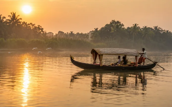 Traditional wooden boat gliding across the calm backwaters during Pathiramanal Island boating in Kerala at sunrise, with coconut palms, misty greenery, and a local boatman.